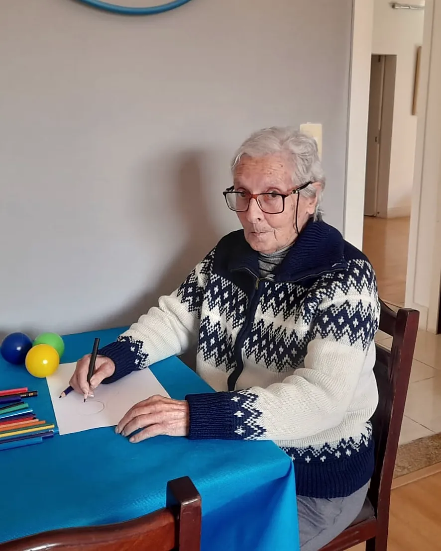 Aluna de cabelos brancos escreve com lápis de cor à mesa, na sala de atividades do Villa Olívias
